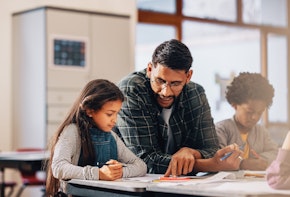 Man mentoring elementary school children. Teacher showing kids how to draw in a class. Male educator assists a young girl on how to complete an art task in a primary class.