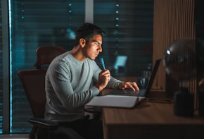 Waist up shot of a young Hispanic male entrepreneur working at night, a laptop and an open writing pad on the desk in front of him. Bright glow from the screen reflecting on his face and in the terrace window. Shutters fully closed in the background.
