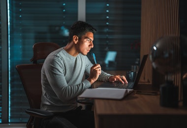 Waist up shot of a young Hispanic male entrepreneur working at night, a laptop and an open writing pad on the desk in front of him. Bright glow from the screen reflecting on his face and in the terrace window. Shutters fully closed in the background.