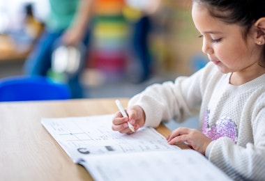 A sweet mixed race elementary student sits at her desk as she works through a writing workbook independently.