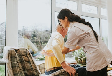 A young female carer helps an elderly male to get dressed by pulling on his jumper, as he sits in an armchair.