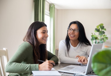 Using their home computer, a mother aids her teenage daughter in completing a virtual high school task.