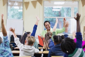 An African-American teacher, a mature woman in her 40s, sitting in front of her class of elementary school students, 6 and 7 years old, reading a book. She has asked a question and most of the children are raising their hands. They are in first grade or second grade.