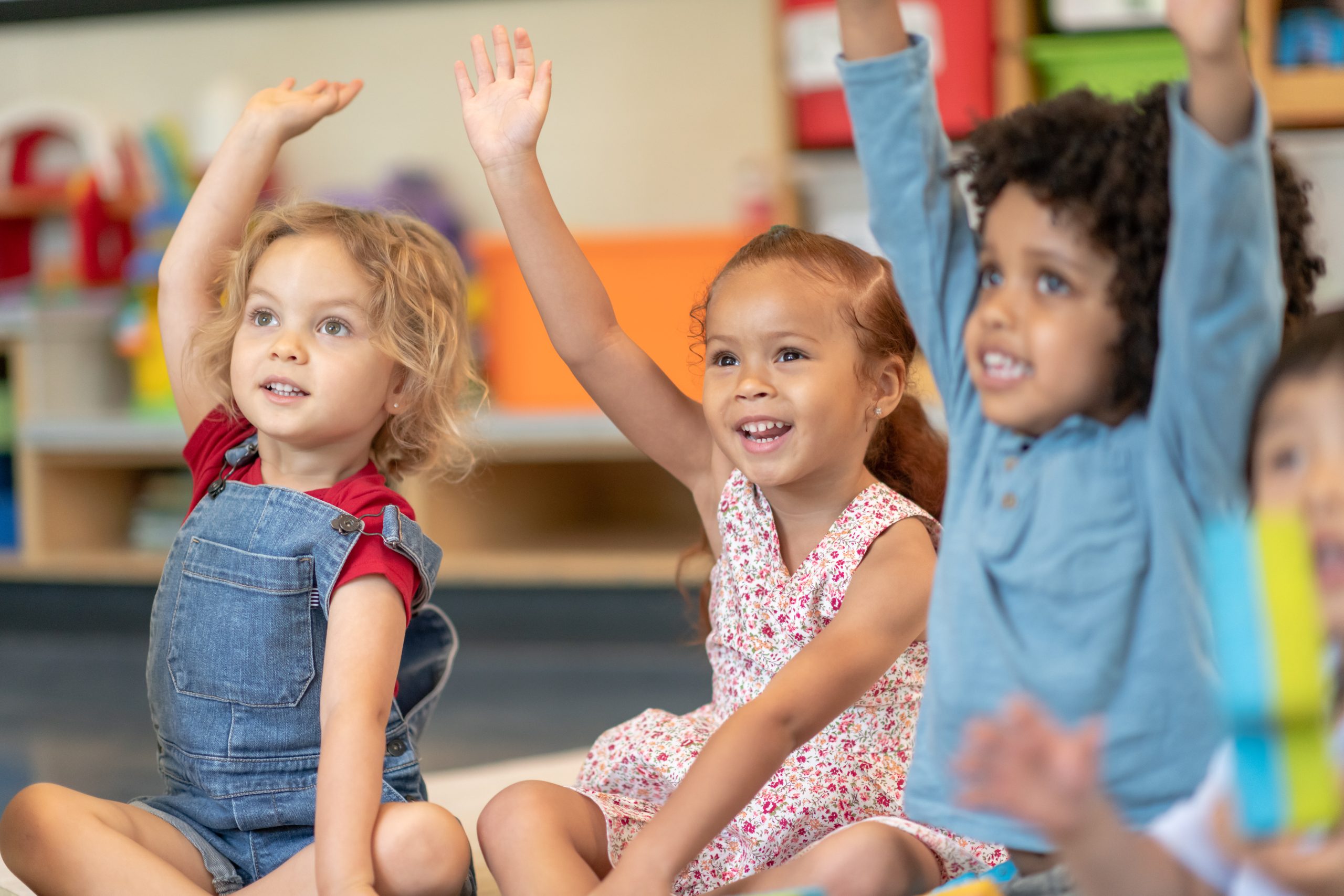 A multi-ethnic group of preschool students is sitting with their legs crossed on the floor in their classroom. The teacher is sitting on the floor facing the children. The happy kids are smiling and listening to the teacher. They have their arms raised and are eager to answer the teacher's question.