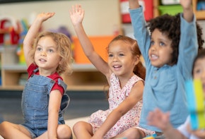 A multi-ethnic group of preschool students is sitting with their legs crossed on the floor in their classroom. The teacher is sitting on the floor facing the children. The happy kids are smiling and listening to the teacher. They have their arms raised and are eager to answer the teacher's question.