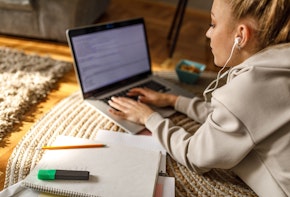 Copy space shot of diligent teenage girl lying on front, on the floor, listening to music via headphones and tying an essay on laptop for homework.