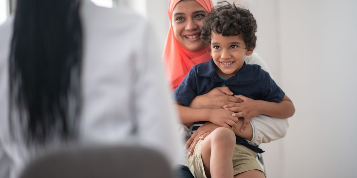 A Muslim mother and son is at the doctor's office together for a check up. The boy is sitting on his mother's lap and she is hugging him tightly.