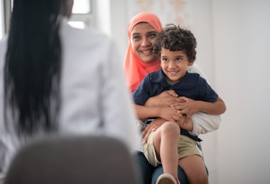 A Muslim mother and son is at the doctor's office together for a check up. The boy is sitting on his mother's lap and she is hugging him tightly.