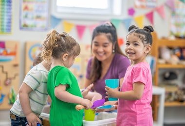 A preschool teacher knees down with a small group of children as they gather around an activity table filled with rice. They are each dressed casually and are smiling as they enjoy playing together.