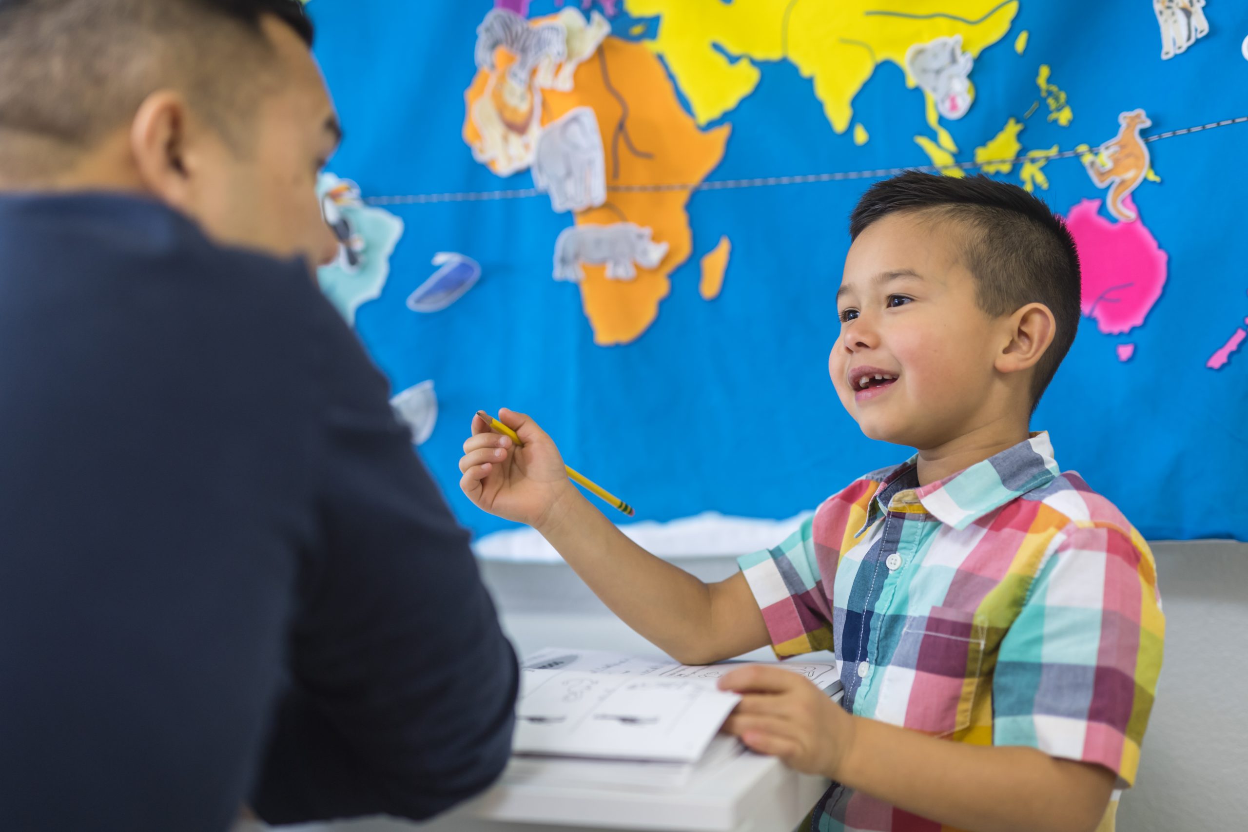 A boy sits at his home desk in front of a colorful world map and writes in his workbook while his teacher - dad! - helps him.