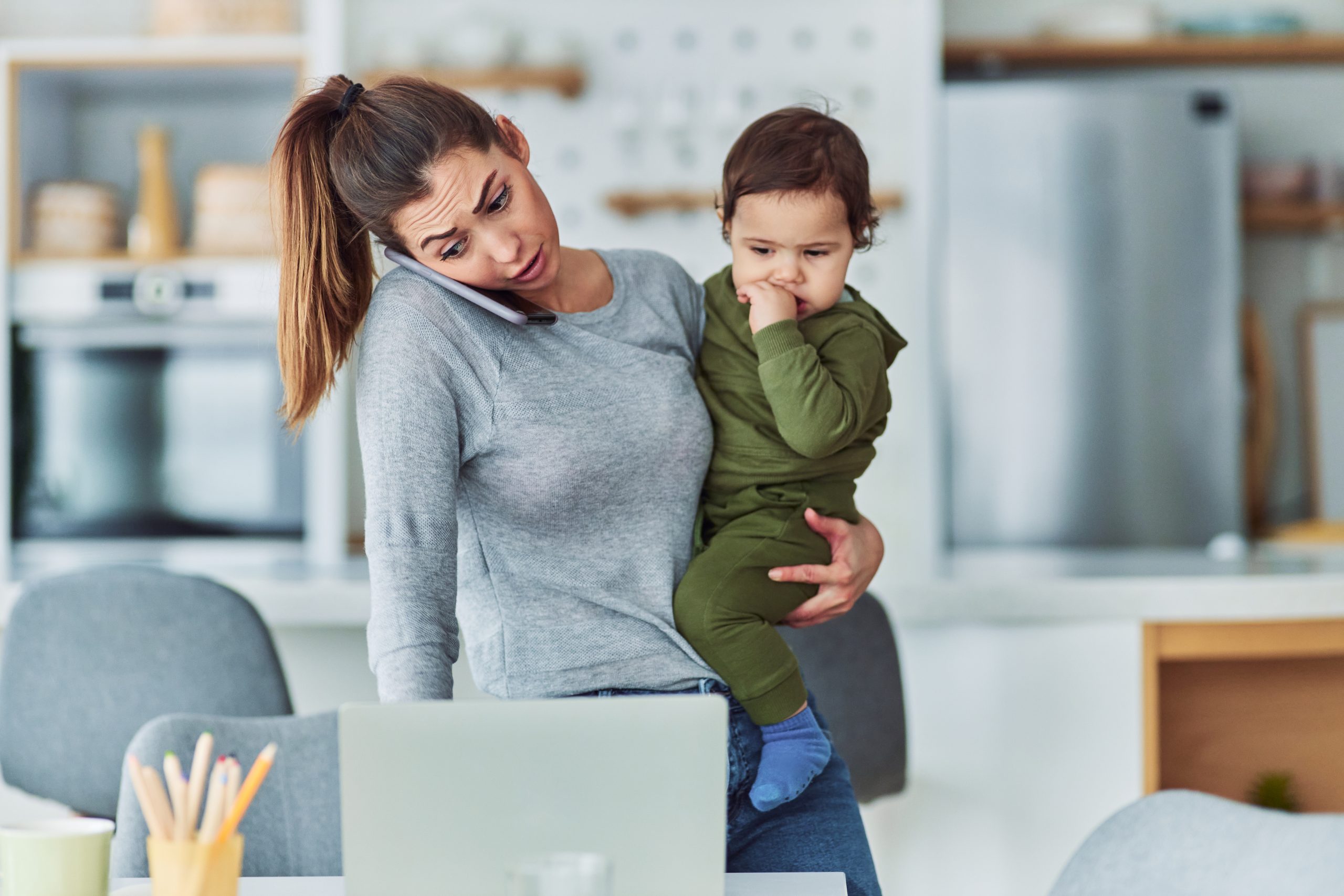 A busy stay-at-home mother multitasking while working on her laptop from home, talking on her phone and carrying her toddler.