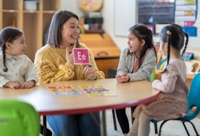 A Kindergarten teacher sits with a small group of students as she teaches them the different sounds each letter of the alphabet makes. Each of the students are dressed casually and paying close attention to their teacher.