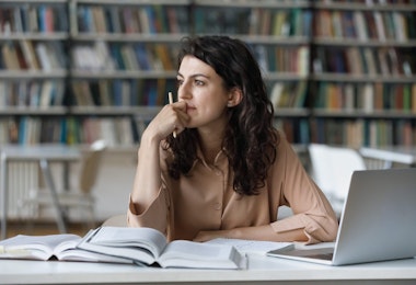 Young student girl sit at table with textbooks and laptop staring aside, studying alone in library, looks pensive and thoughtful search solution, prepare for exam, makes task feels confused or puzzled