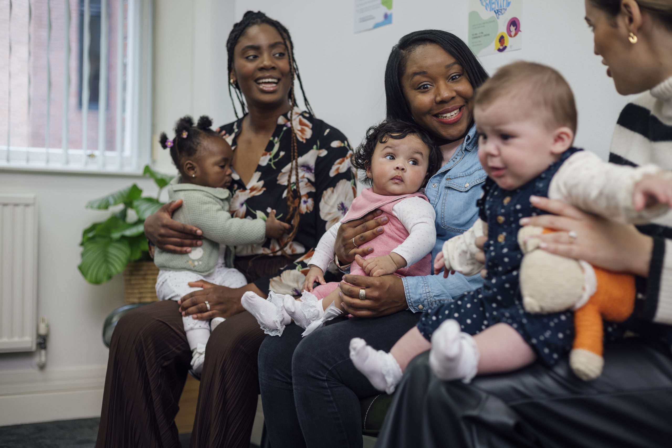 A shot of a small group of female adults sitting together with their babies in the waiting room of a baby clinic in Newcastle-upon-Tyne, North East England. They are talking and smiling, their babies distracted by what's going on around them. One baby is crying.