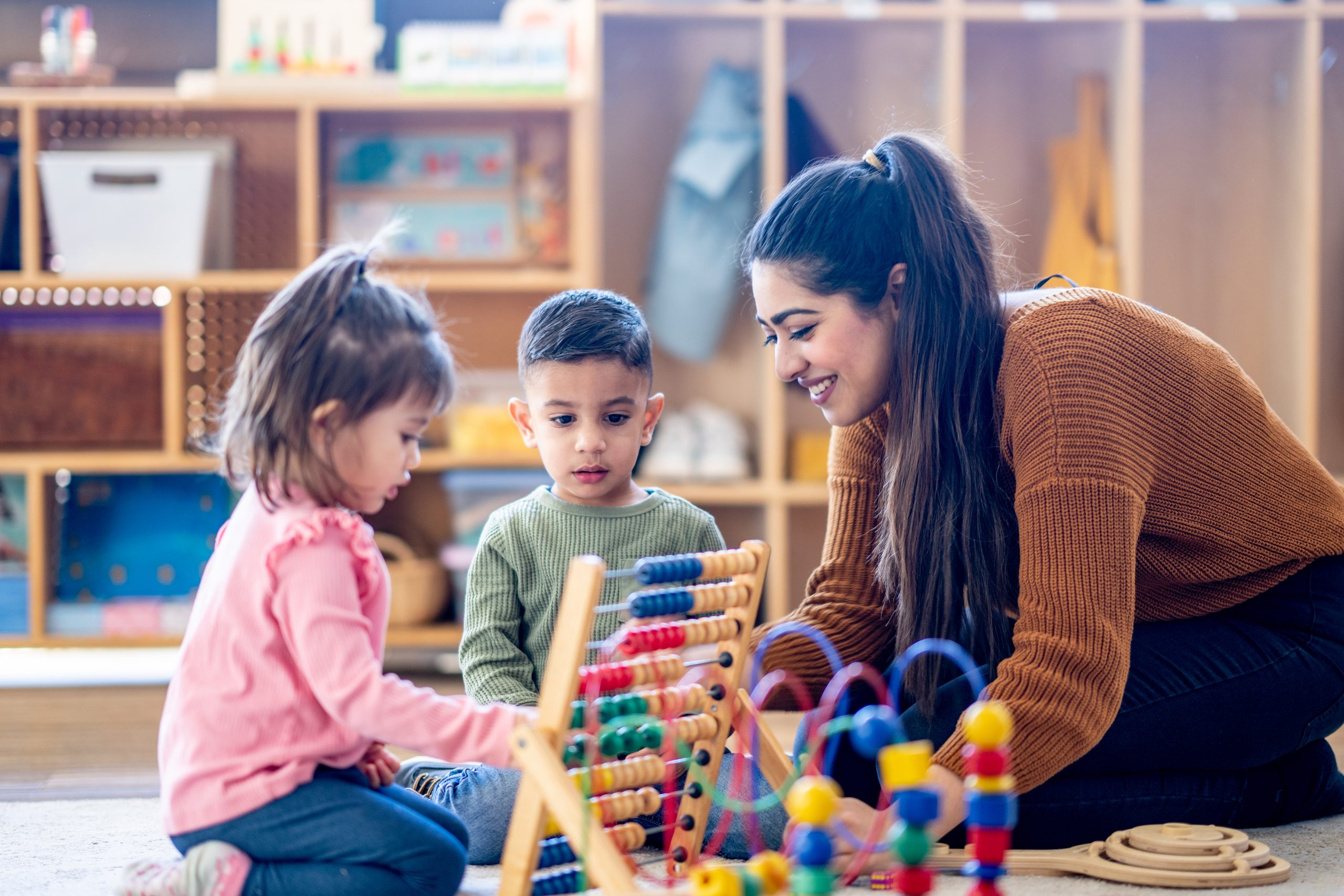 A female teacher of Middle Eastern decent sits on the floor with a small group of students as they learn to count together with an abacus.  They are each dressed casually and have neutral expressions on their faces as they work together.