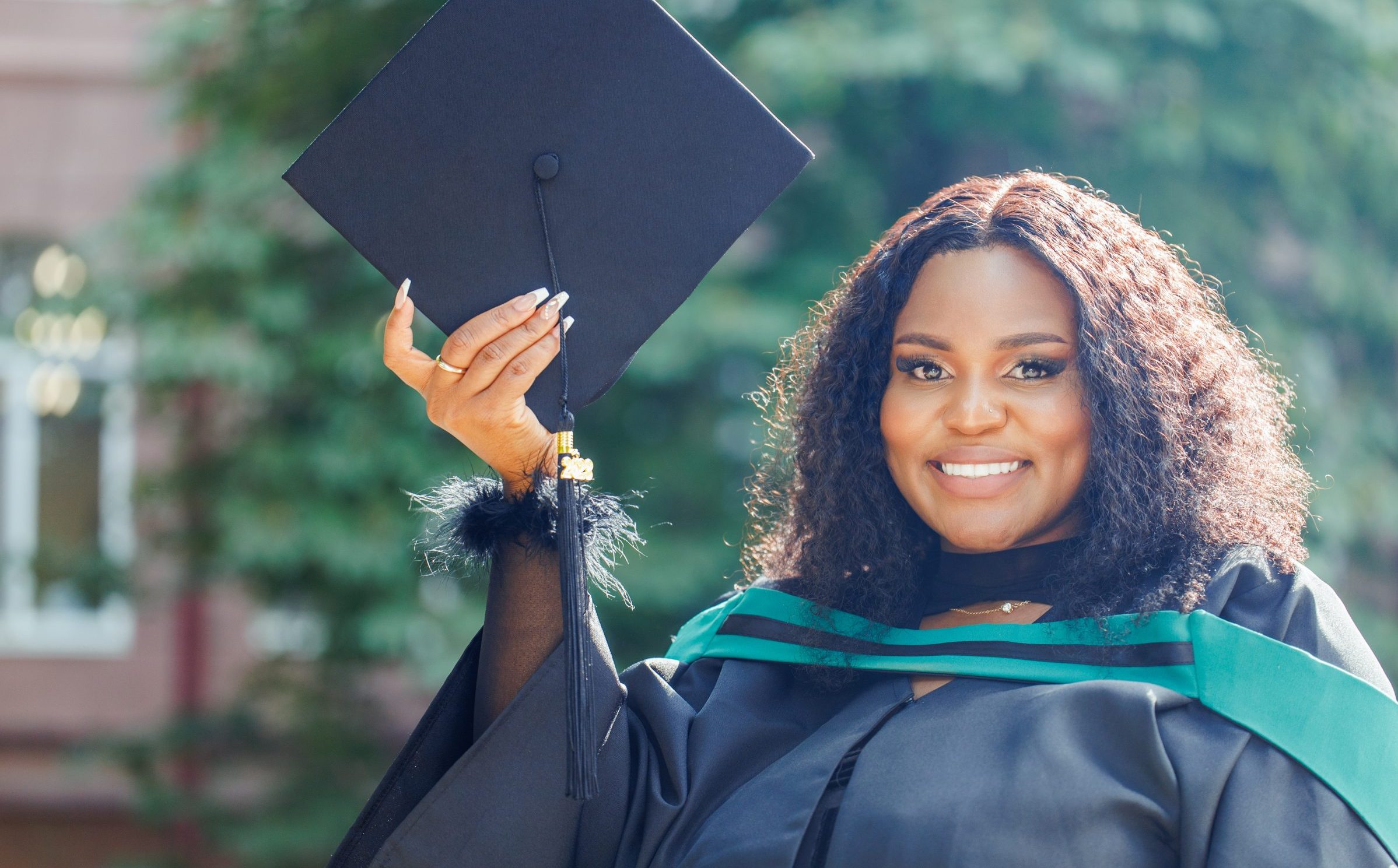 Pursuing dreams: Happy young black female graduate with natural curly black hair in graduation gown holding mortarboard in one hand, showing it to camera and smiling, standing in green space in university courtyard