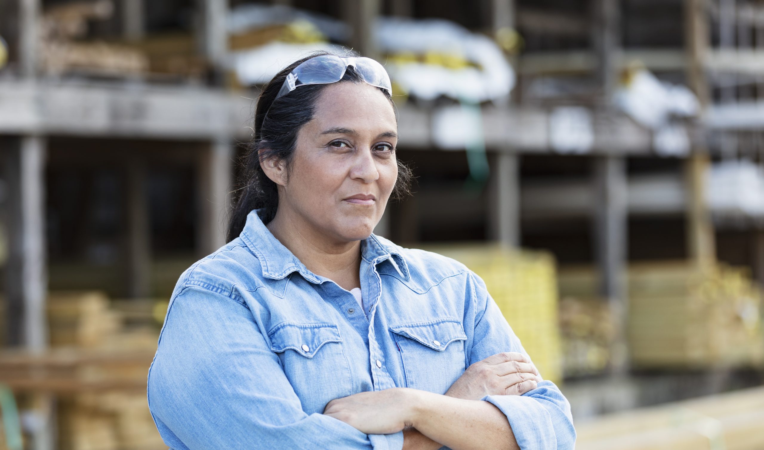A mature Hispanic woman working in a lumberyard or building supply store, looking at the camera with her arms crossed. Shelves stacked with lumber are out of focus in the background.