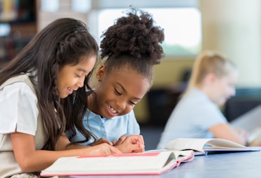 Happy female elementary age students in school uniforms are sitting at a desk in the school library and reading a book together. A Filipino elementary age girl sits with an African American classmate and reads a book together in the school library.