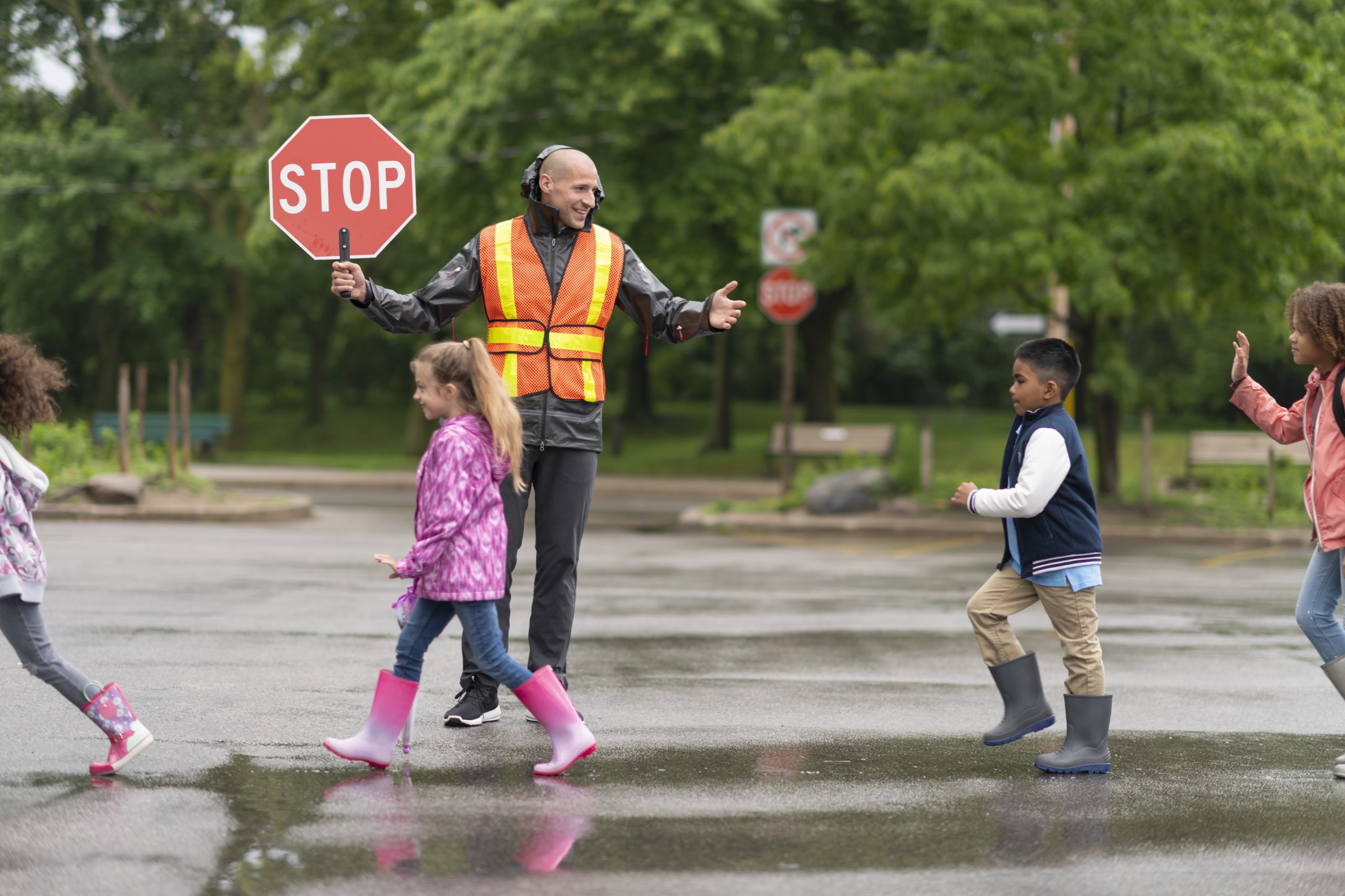 A multi-ethnic group of elementary students are on their way to school. The children are crossing the street with the assistance of a smiling crossing guard. The male crossing guard is happy to help the kids stay safe. It's a rainy day and the children are wearing rain coats and rain boots. The crossing guard is holding a stop sign and his wearing a reflective safety vest.