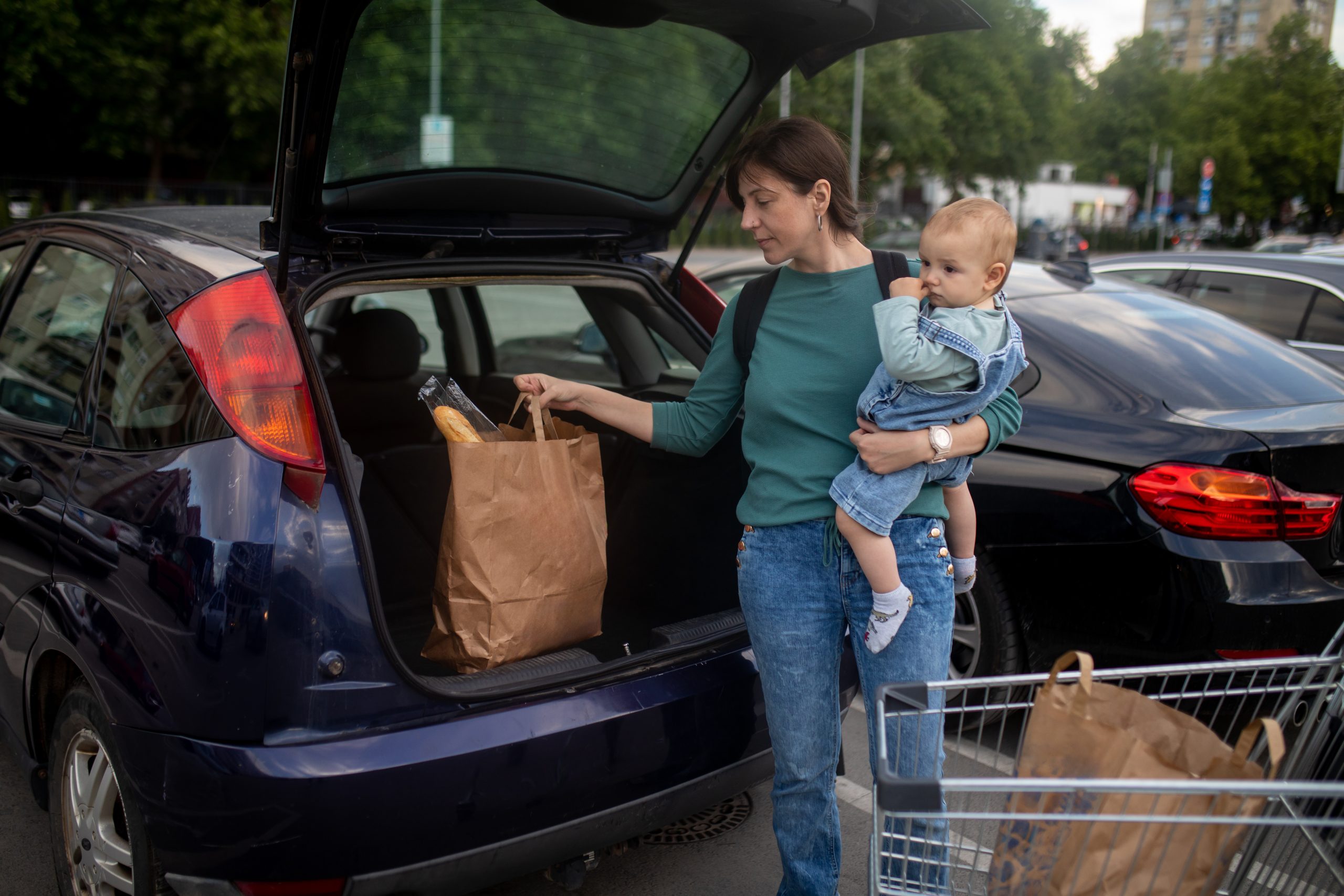 Young woman, with brown hair, has finished grocery shopping in the supermarket. She is on the parking and holding her son, cute baby boy, while she packing a bag of groceries into the trunk of her car.