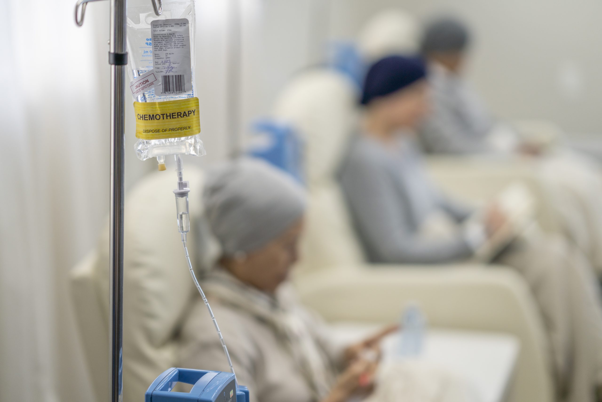 Three women sit in individual chairs as they receive their Chemotherapy by intravenous.  They are each dressed comfortably and have head scarves on to keep them warm as they engage in the activity they each brought to keep them occupied.