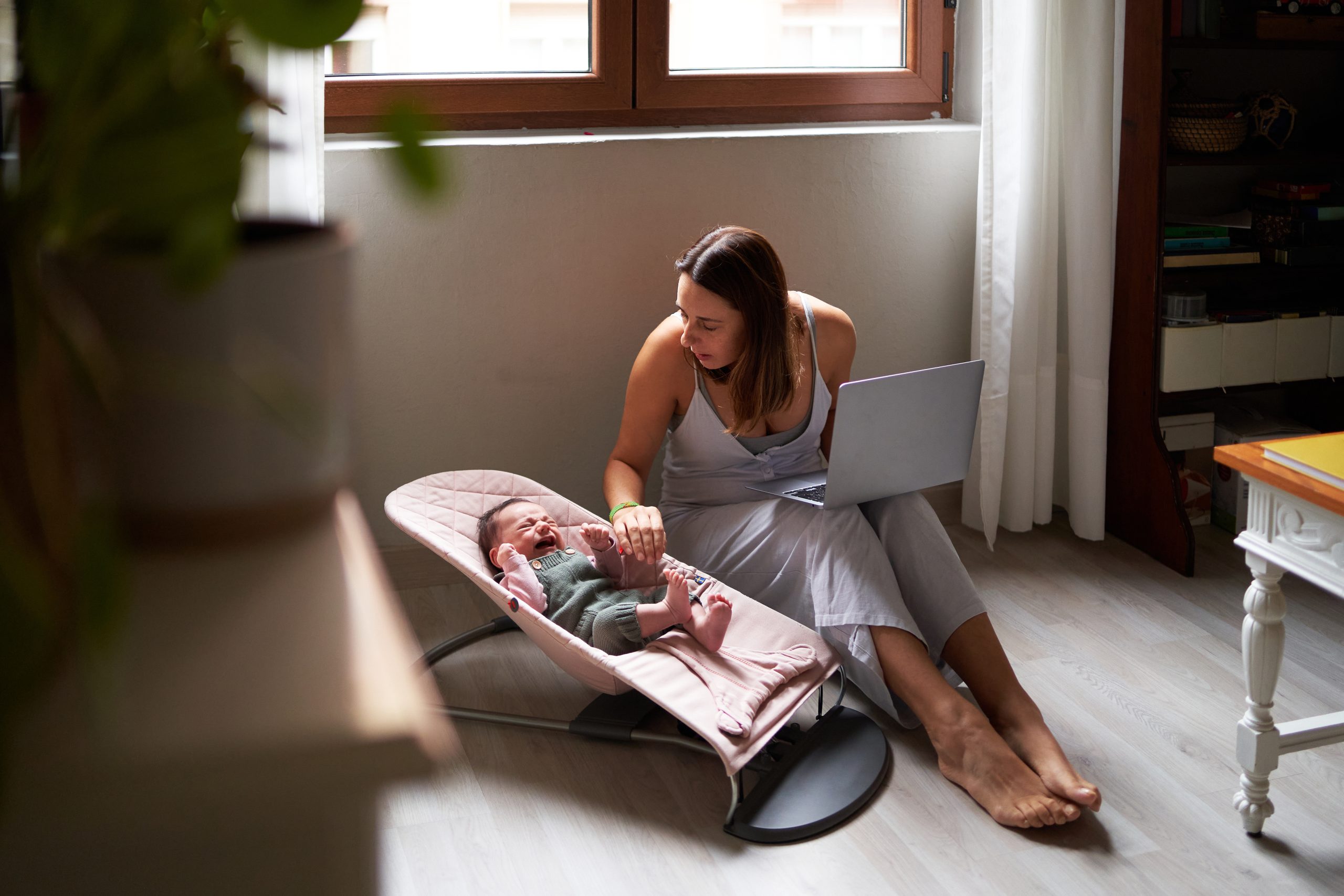 Mother using a laptop while consoling a baby lying on a baby swing at home