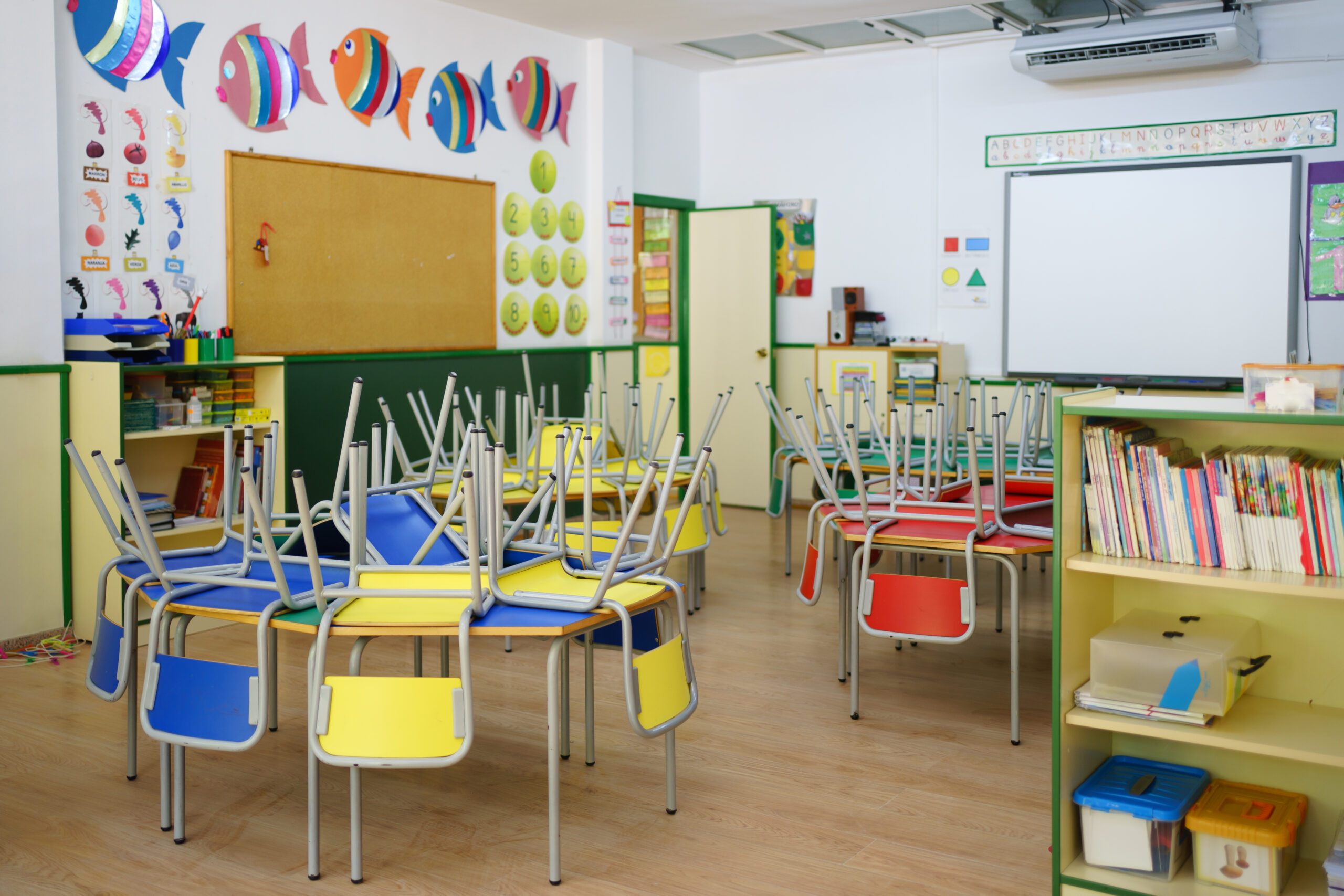 Wide view of a children's classroom at the school. Red, yellow and blue chairs arranged on the tables, white board and other school equipment.