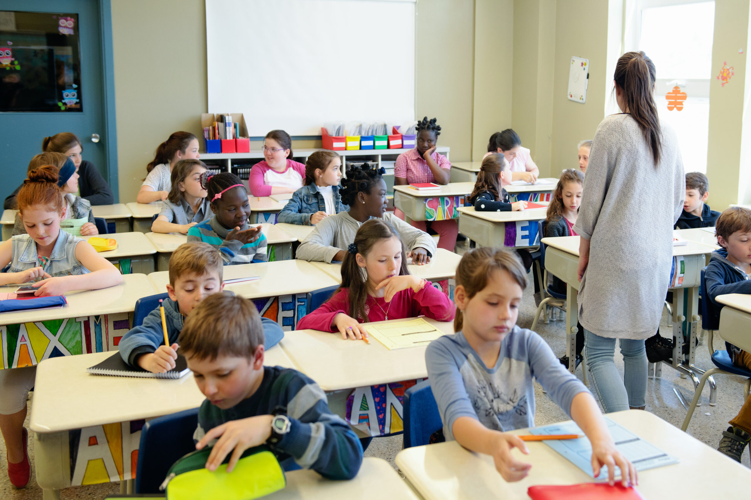 Multi-ethnic schoolgirls and shcoolboys sit into the class for their first day at school. Some of them are shy and other expressive.  There is multicolour name in the front of the desk. Photo was taken in Quebec Canada.