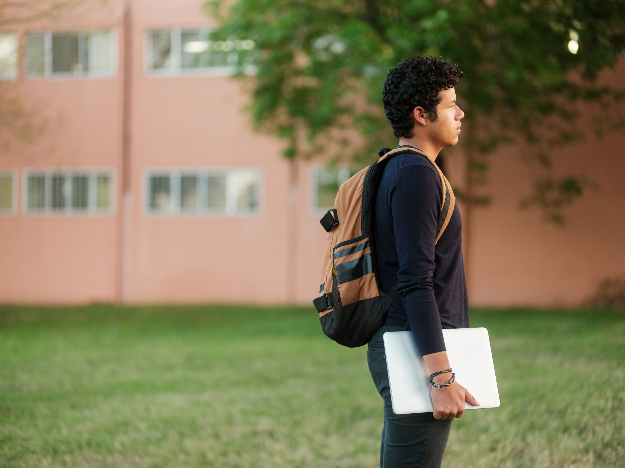 Side view of a male college student standing by himself outside holding a laptop and carrying a backpack.