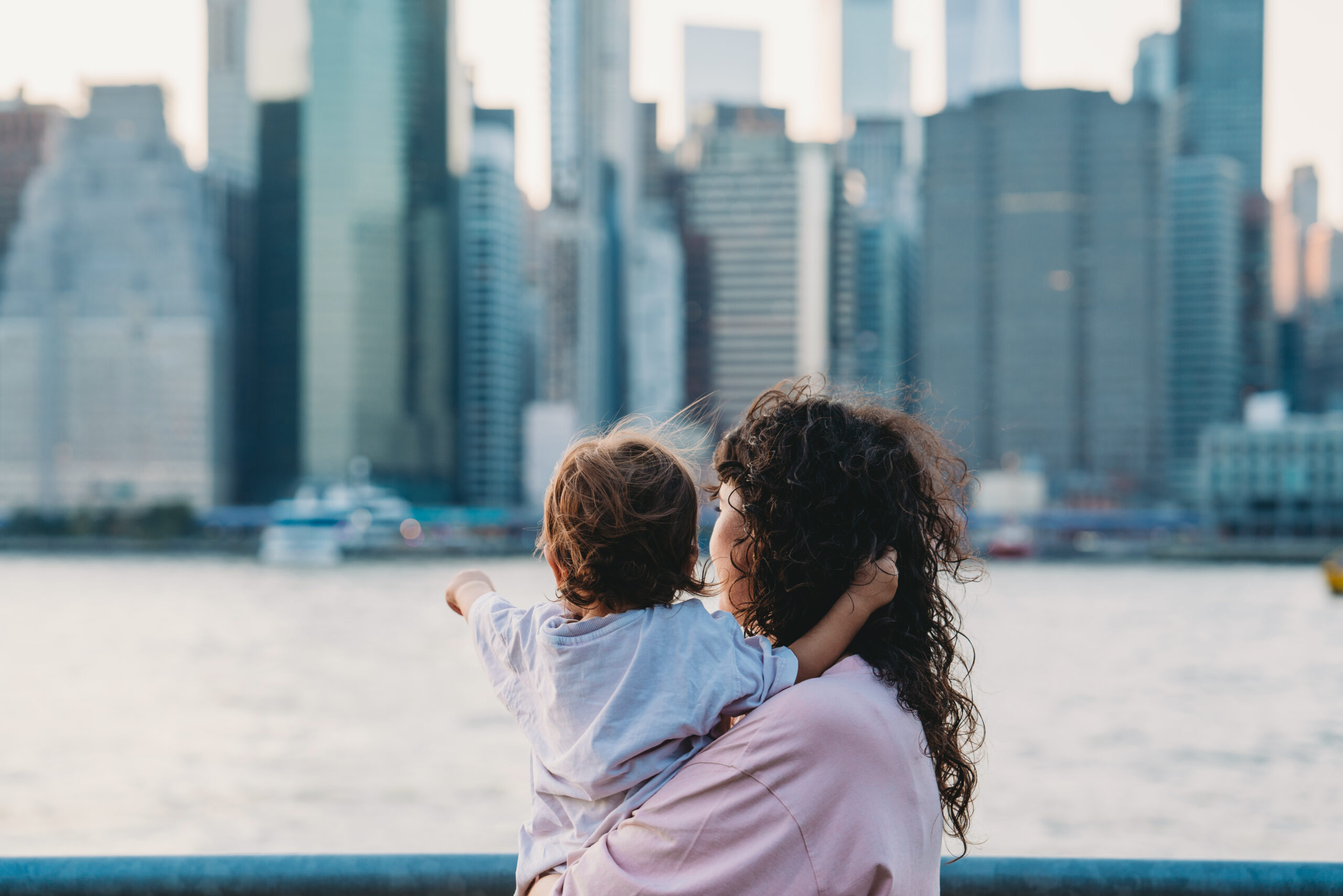 Mother is holding her baby while pointing at the manhattan skyline from across the east river