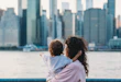Mother is holding her baby while pointing at the manhattan skyline from across the east river