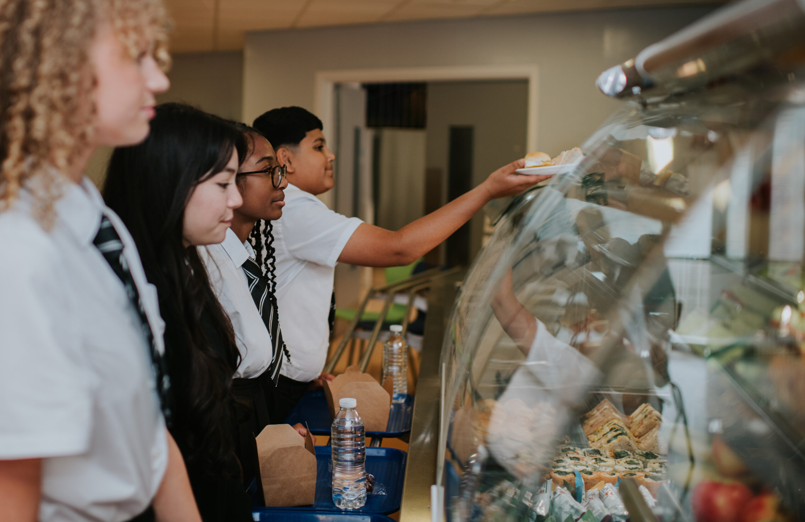 A group of secondary school children wearing uniforms wait in line wearing uniforms. They make their food choices in the school cafeteria. Focus is on a young lad reaching up for a tray.