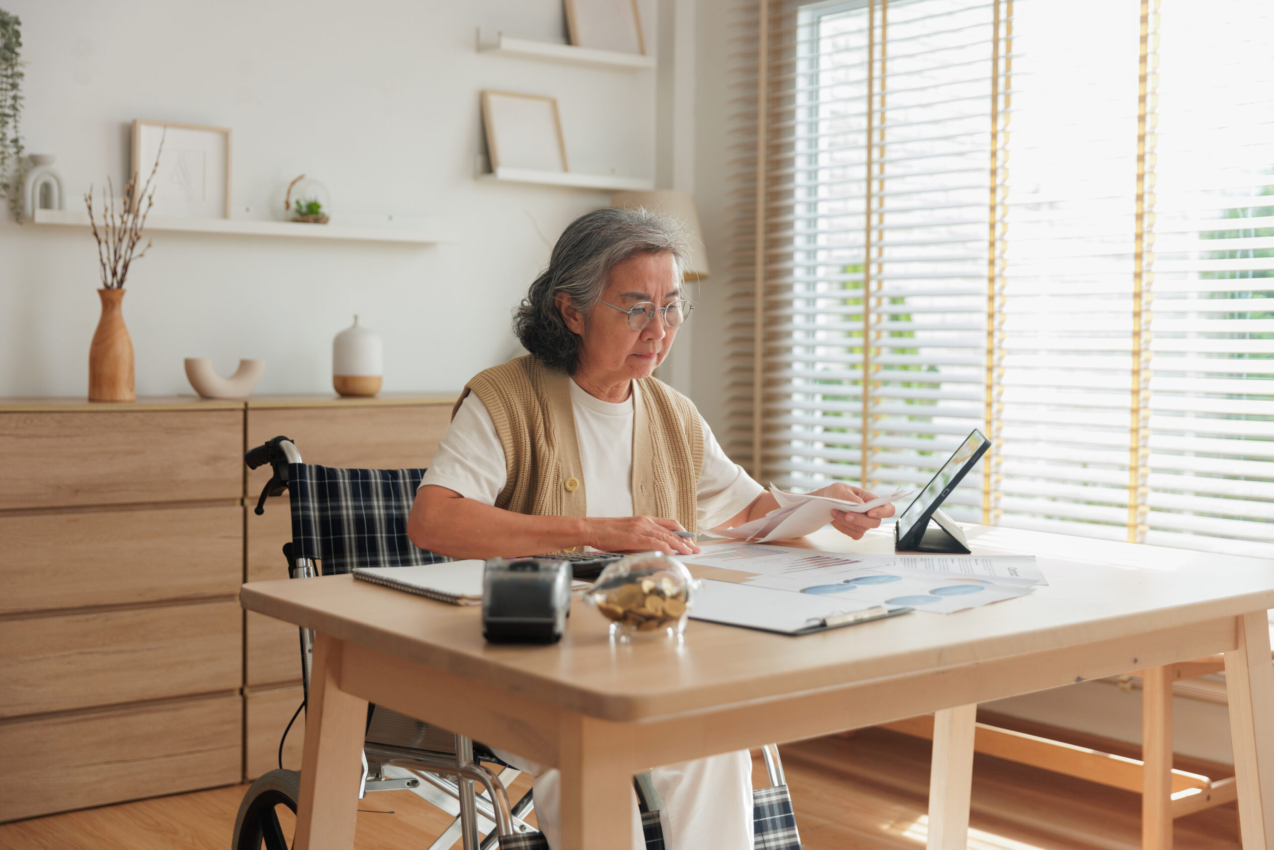 Portrait of an elderly Asian woman with a disability sitting in a wheelchair at a table. She is stressed while calculating expenses, holding paper bills, and checking her savings with a piggy bank and tablet. Concept of retirement costs, inflation, and pension management.