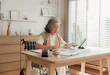 Portrait of an elderly Asian woman with a disability sitting in a wheelchair at a table. She is stressed while calculating expenses, holding paper bills, and checking her savings with a piggy bank and tablet. Concept of retirement costs, inflation, and pension management.